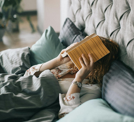 Eine Frau liegt müde im Bett und hält sich ein Buch über ihr Gesicht